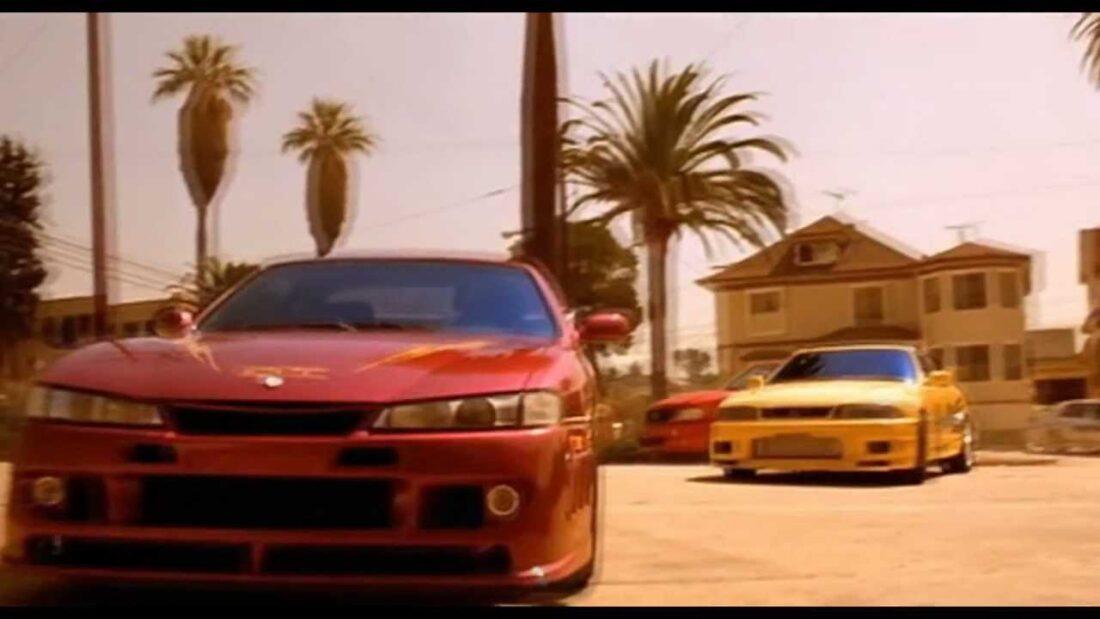 Two sports cars, one red and one yellow, parked on a street with palm trees in the background.